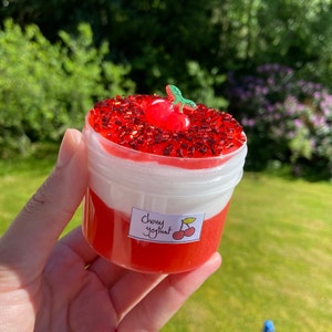 Puede incluir: Un recipiente de pl&aacute;stico transparente lleno de slime rojo con sabor a cereza cubierto con sprinkles rojos y una peque&ntilde;a cereza roja con un tallo verde. El recipiente tiene una etiqueta blanca que dice "Cherry Yogurt".
