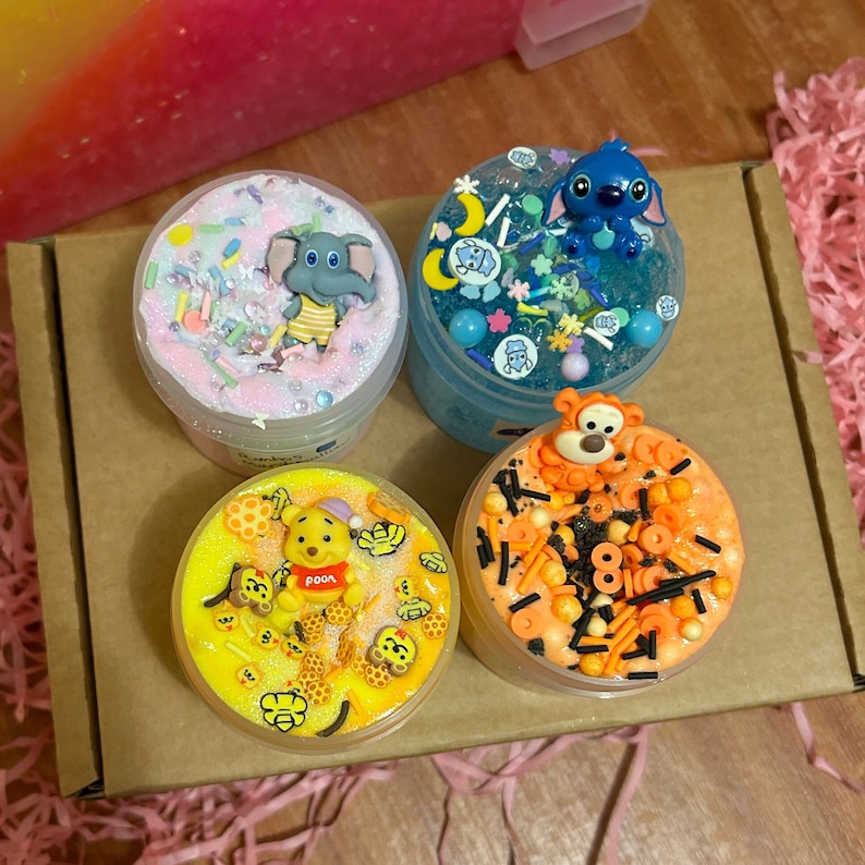 May include: Four clear plastic containers filled with colorful slime and topped with various cartoon character figurines. The slime is decorated with sprinkles, glitter, and other embellishments. The characters include Winnie the Pooh, Tigger, Stitch, and Dumbo.