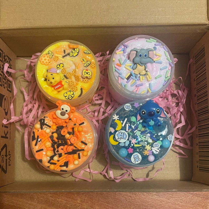 May include: Four small jars of slime with different colors and decorations. The slime is in a box with pink shredded paper. The slime jars have a Winnie the Pooh character, a Tigger character, a Stitch character, and an elephant character on top.