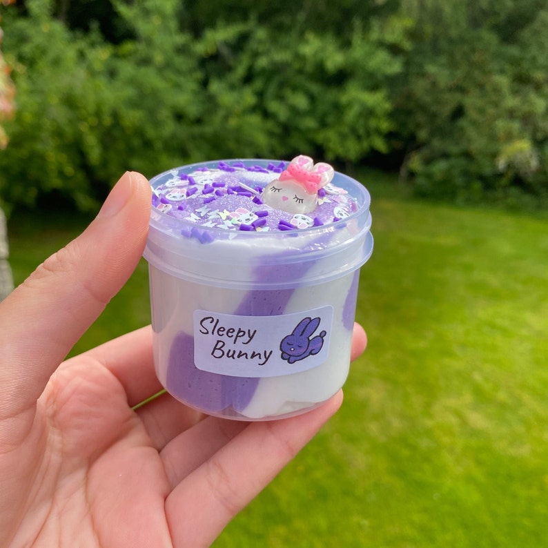 May include: Purple and white slime in a clear plastic container with a white label that says "Sleepy Bunny". A small white bunny with pink ears and a pink bow is on top of the slime.