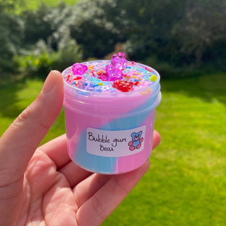 May include: A clear plastic container filled with pink and blue slime with a swirl design. The slime is topped with colorful sprinkles, gummy bears, and a label that reads "Bubble gum Bear".