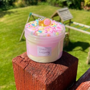 May include: A clear plastic container filled with white and pink slime with colorful sprinkles and a small snail figurine on top. The container has a label that says "Snail Frosting".