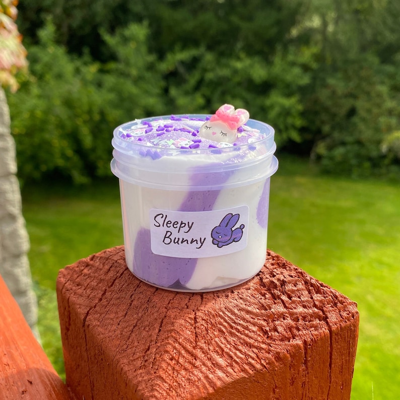 May include: A clear plastic container filled with purple and white slime with a small white bunny charm on top. The container has a label that says "Sleepy Bunny".