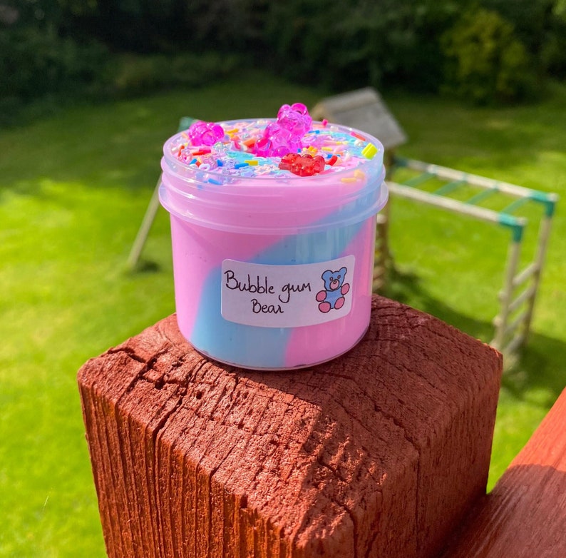 May include: A clear plastic jar filled with pink and blue slime with a swirl design. The slime is topped with colorful sprinkles and gummy bears. The jar has a label that says "Bubble gum Bear".