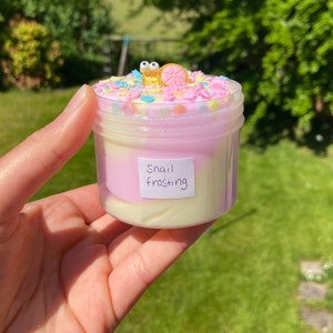 May include: A clear jar filled with white and pink slime topped with colorful sprinkles and a small snail charm. The jar has a label that reads "Snail Frosting".
