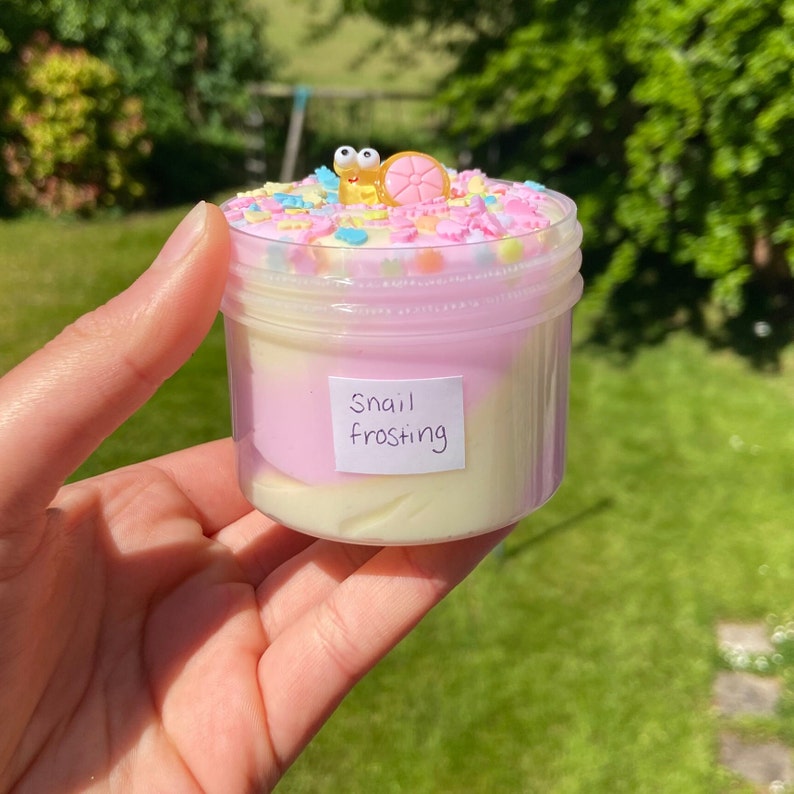 May include: A clear jar filled with white and pink slime topped with colorful sprinkles and a small snail charm. The jar has a label that reads "Snail Frosting".