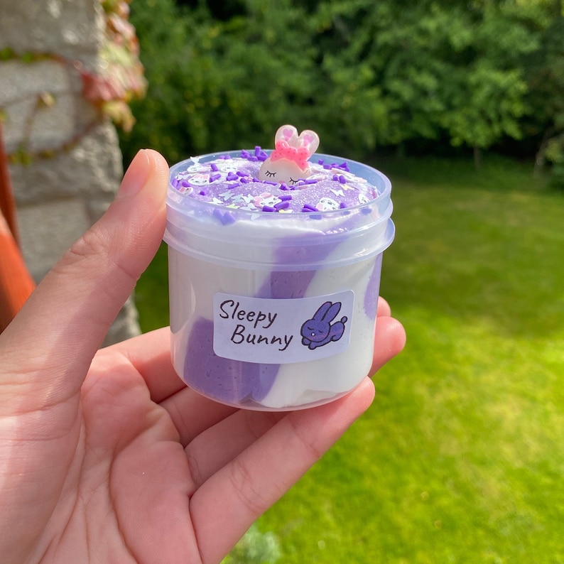 May include: Purple and white slime in a clear plastic container with a white label that says "Sleepy Bunny". A small pink bunny charm is on top of the slime.