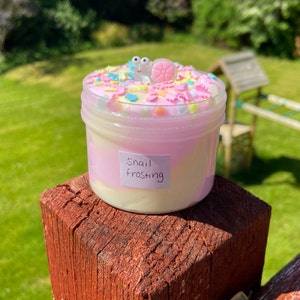 May include: A pink and white slime with a snail charm and colorful sprinkles. The slime is in a clear plastic container with a label that says "Snail Frosting".