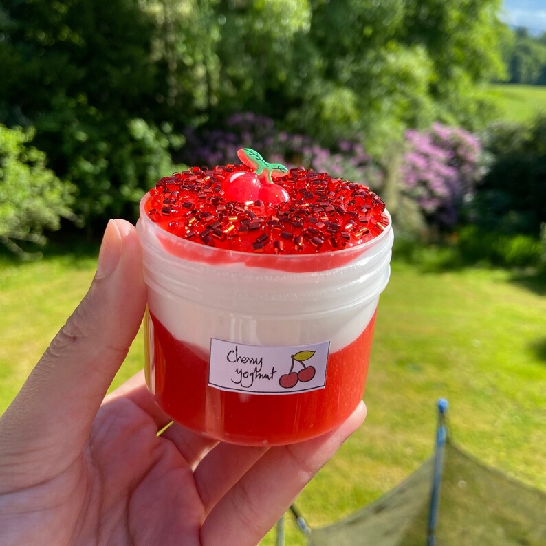 Puede incluir: Un tarro de pl&aacute;stico transparente lleno de slime rojo y blanco. El slime est&aacute; cubierto de purpurina roja y una cereza roja. El tarro tiene una etiqueta que dice "Yogurt de cereza".