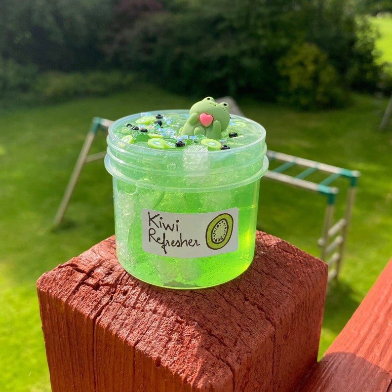 Pode incluir: Um frasco transparente de slime verde com um adesivo de fatia de kiwi e o texto "Kiwi Refresher". Uma pequena figura de sapo verde est&aacute; em cima do slime.