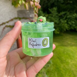 Pode incluir: Um frasco de slime verde transparente com um amuleto de sapo verde no topo. O frasco tem um r&oacute;tulo branco que diz "Kiwi Refresher" com uma ilustra&ccedil;&atilde;o de kiwi verde.