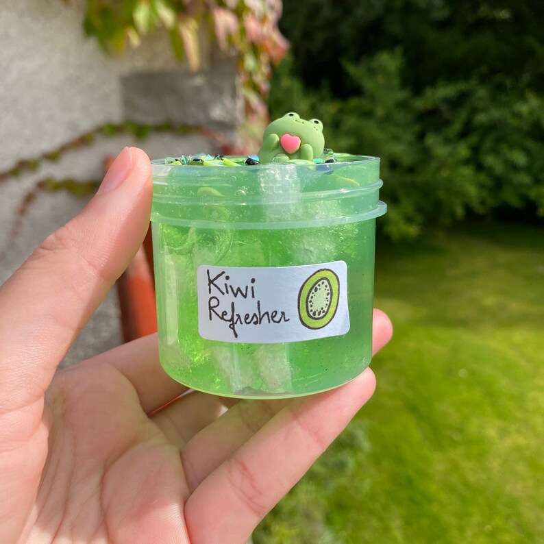 Pode incluir: Um frasco de slime verde transparente com um amuleto de sapo verde no topo. O frasco tem um r&oacute;tulo branco que diz "Kiwi Refresher" com uma ilustra&ccedil;&atilde;o de kiwi verde.