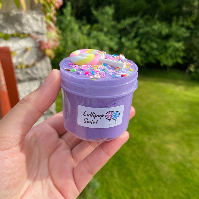 May include: Purple slime with colorful sprinkles and a lollipop on top. The slime is in a clear plastic container with a label that says "Lollipop Swirl".