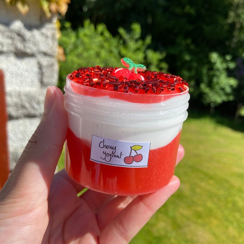 Puede incluir: Un recipiente de pl&aacute;stico transparente lleno de slime rojo y blanco, cubierto con purpurina roja y una peque&ntilde;a cereza de pl&aacute;stico. El recipiente tiene una etiqueta que dice "Cherry Yogurt".