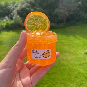 May include: Orange-coloured slime in a clear jar with a label that says "Fanta Crush". An orange slice is on top of the jar.
