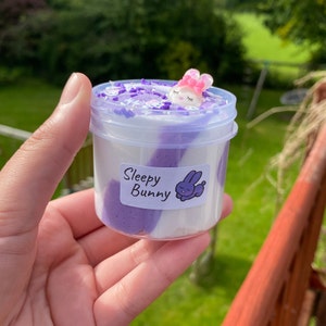 May include: A clear plastic container filled with purple and white slime. The slime has a swirl pattern and is topped with white and purple sprinkles. A small white bunny charm with pink ears is resting on top of the slime. The container has a label that reads "Sleepy Bunny".