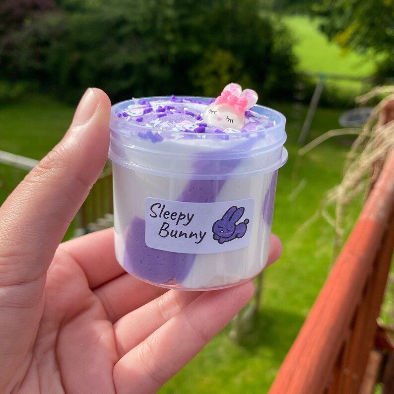 May include: A clear plastic container filled with purple and white slime. The slime has a swirl pattern and is topped with white and purple sprinkles. A small white bunny charm with pink ears is resting on top of the slime. The container has a label that reads "Sleepy Bunny".