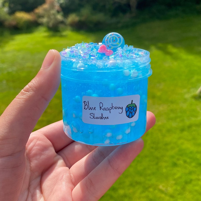 Puede incluir: Un peque&ntilde;o frasco transparente lleno de slime azul con purpurina blanca e iridiscente. El frasco tiene una etiqueta que dice "Blue Raspberry Slushue" con un ar&aacute;ndano de dibujos animados.