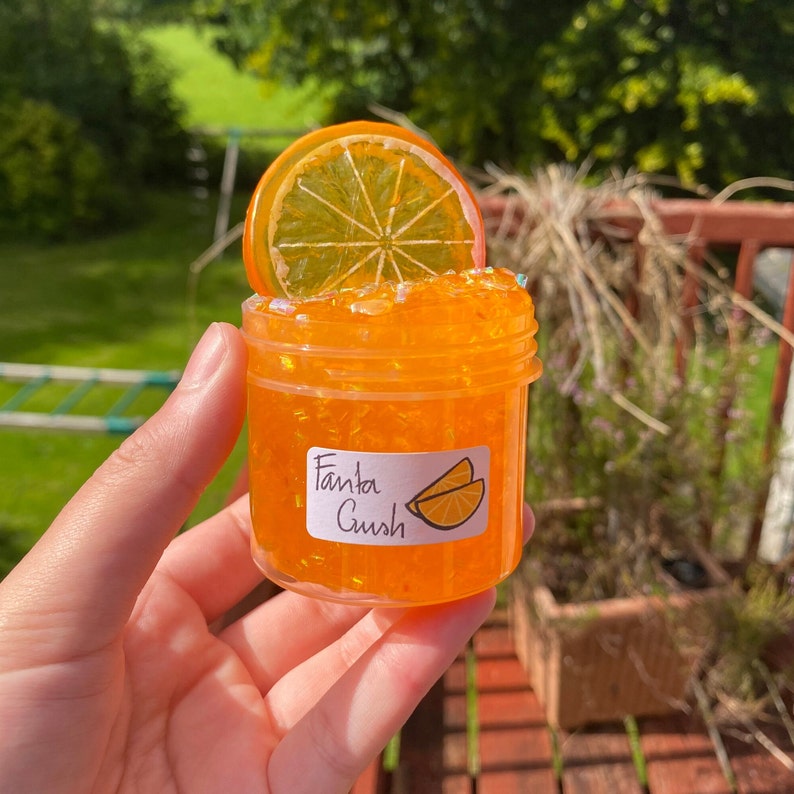 May include: A clear jar filled with orange slime with a slice of orange on top. The jar has a label that says "Fanta Gush".