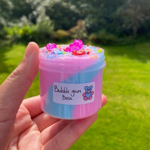 May include: A clear plastic container filled with pink and blue slime with a swirl design. The slime is topped with colorful sprinkles and gummy bears. The container has a white label that says "Bubble gum Bear".