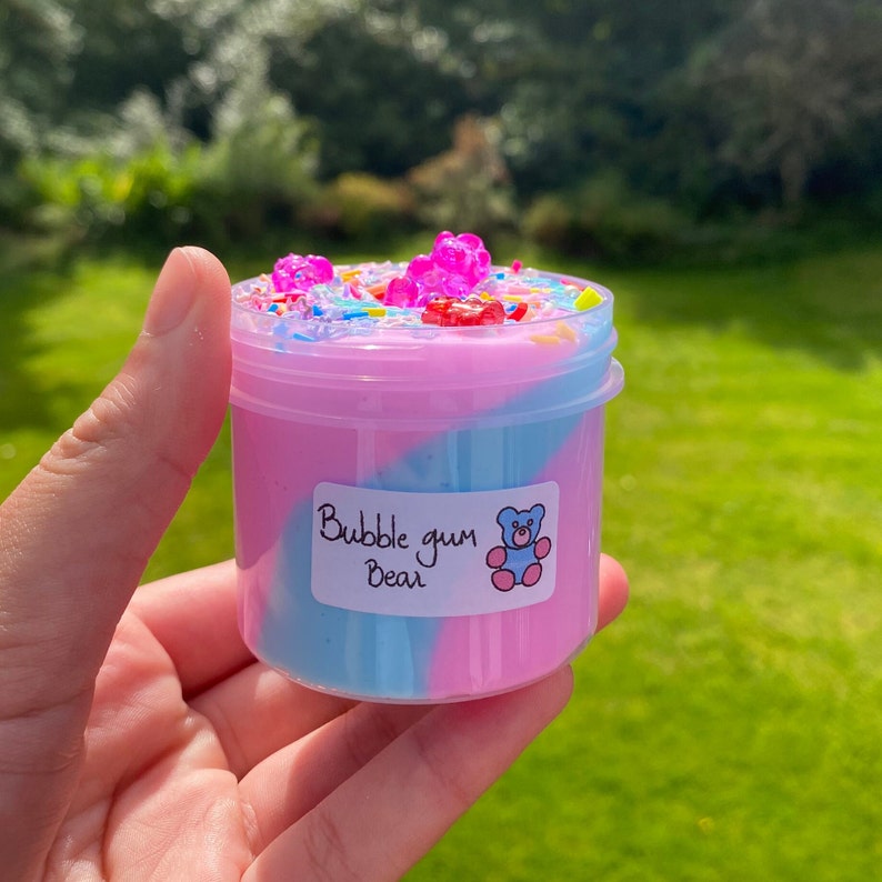 May include: A clear plastic container filled with pink and blue slime with a swirl design. The slime is topped with colorful sprinkles and gummy bears. The container has a white label that says "Bubble gum Bear".