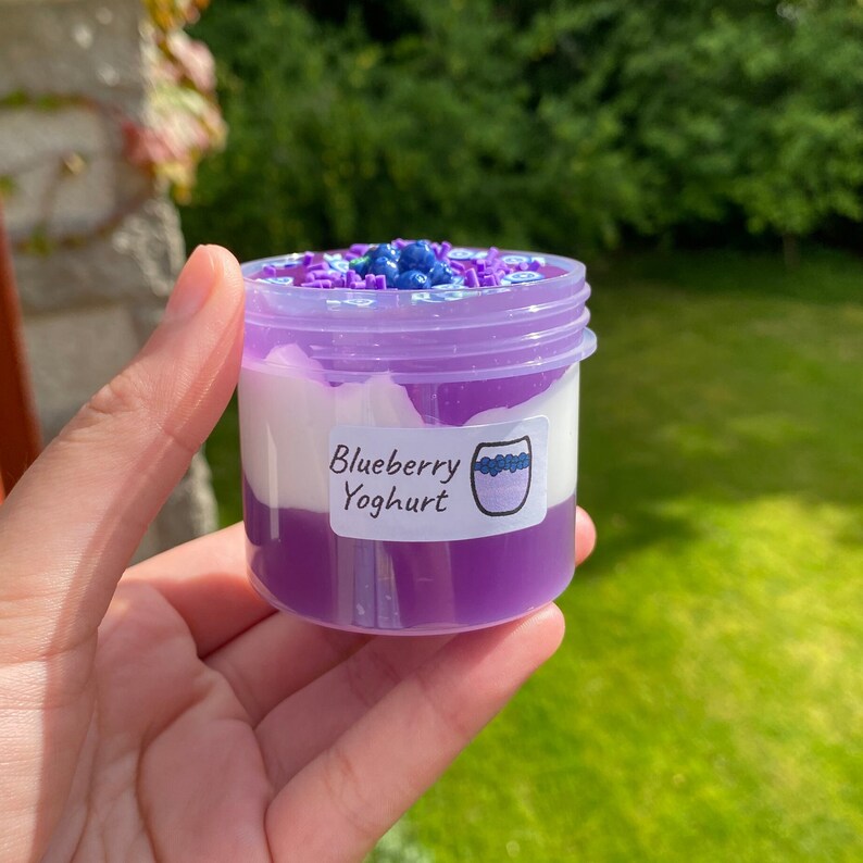 May include: A small purple jar with a white lid filled with a purple and white slime. The jar has a label that says "Blueberry Yoghurt".