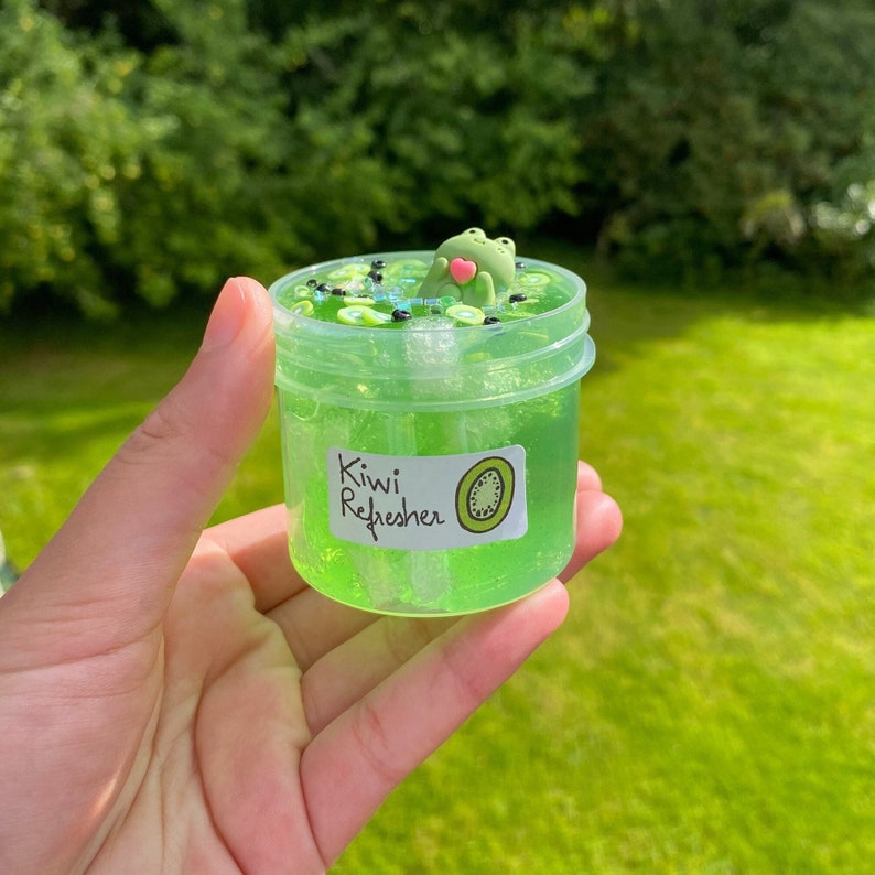 Pode incluir: Um frasco transparente cheio de slime verde, fatias de kiwi e contas pretas. Um pequeno amuleto de sapo verde flutua no topo. O frasco tem um r&oacute;tulo que diz "Kiwi Refresher".