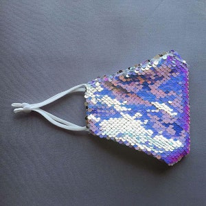 May include: A white face mask with iridescent sequins in shades of purple, pink, and blue. The mask has white elastic straps.