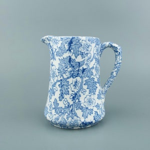 May include: A small, white ceramic jug with a blue floral pattern. The jug has a curved handle and a spout for pouring. The floral design features leaves and flowers. The jug is likely used for serving liquids.