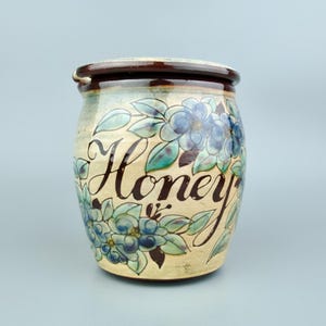 May include: A ceramic honey pot with a lid. The pot is beige with blue floral designs and green leaves. The word "Honey" is written in a dark brown script across the front. The rim and lid are dark brown.