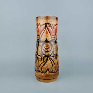 May include: A tall, ceramic vase with a cylindrical shape. The vase features a brown and tan speckled background with a repeating pattern of black and red floral designs. The vase has a wide base and a narrow opening.