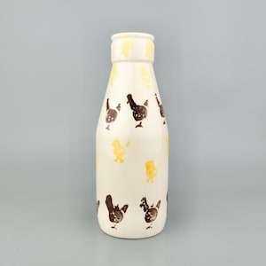 May include: A cream-coloured ceramic bottle decorated with brown chicken and yellow chick motifs. The bottle has a narrow neck and a rounded body. The design is simple and rustic, suitable for farmhouse decor.