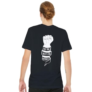 May include: Black t-shirt featuring a white clenched fist graphic with a banner stating "WILL TRADE RACISTS FOR REFUGEES". The shirt is made of a soft material.