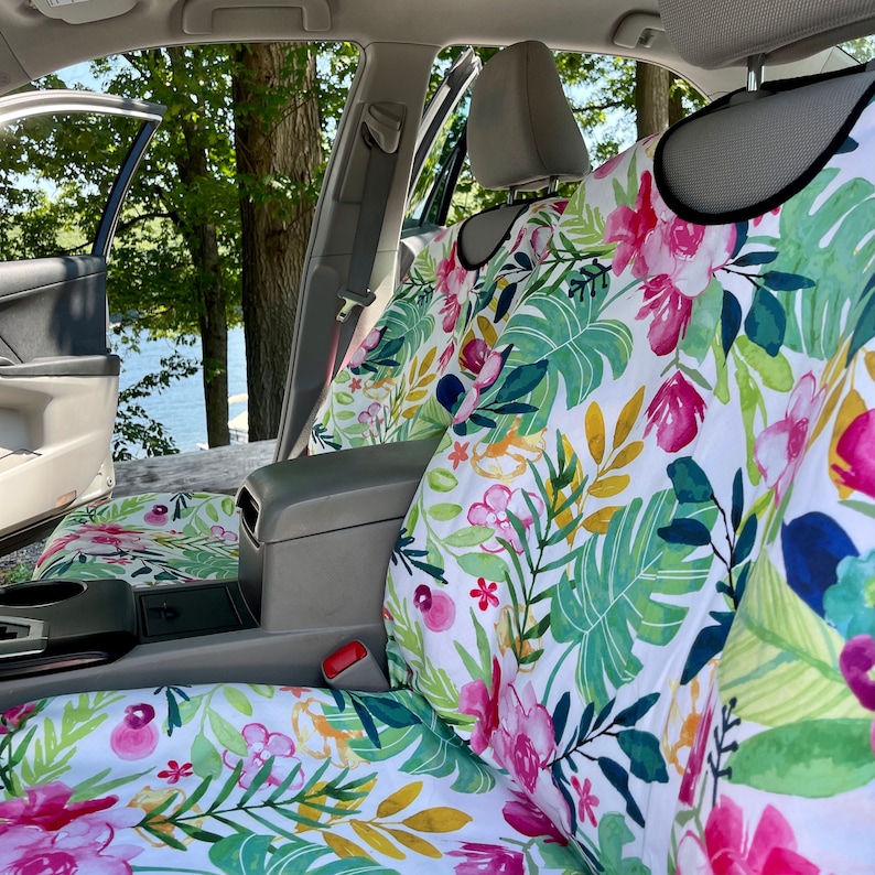 May include: A car seat cover set featuring a white background with a vibrant watercolor print of pink flowers, green tropical leaves, and yellow foliage. The covers are fitted over the front and back seats of a vehicle with a gray interior.