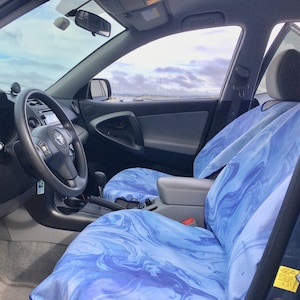 May include: A car interior featuring front seats covered in a blue and white marbled pattern fabric. The seats have a smooth, fluid design with varying shades of light and dark blue swirls. The vehicle includes a black steering wheel and dashboard.