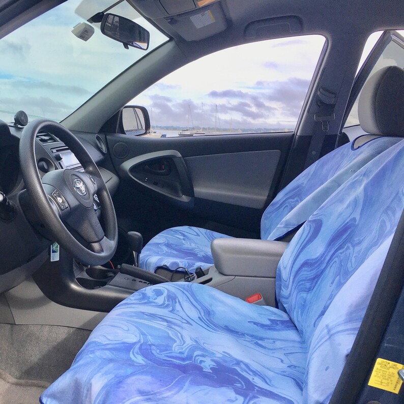 May include: A car interior featuring front seats covered in a blue and white marbled pattern fabric. The seats have a smooth, fluid design with varying shades of light and dark blue swirls. The vehicle includes a black steering wheel and dashboard.