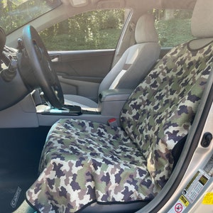 May include: A camouflage print car seat cover featuring a pattern of green, brown, and black shapes. The fabric cover is draped over the front passenger seat of a vehicle, extending from the headrest down to the seat cushion with black trim.