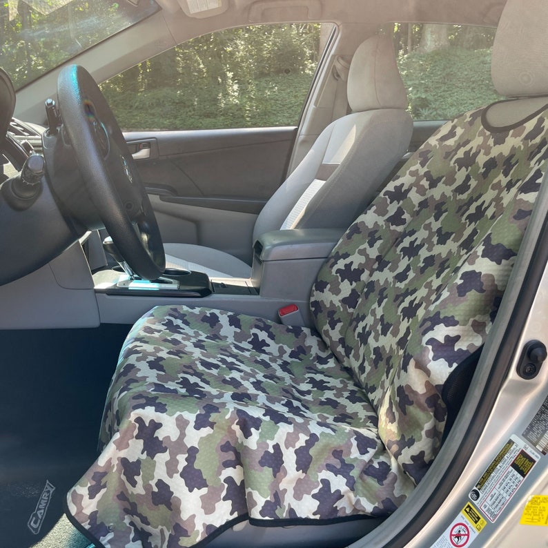 May include: A camouflage print car seat cover featuring a pattern of green, brown, and black shapes. The fabric cover is draped over the front passenger seat of a vehicle, extending from the headrest down to the seat cushion with black trim.