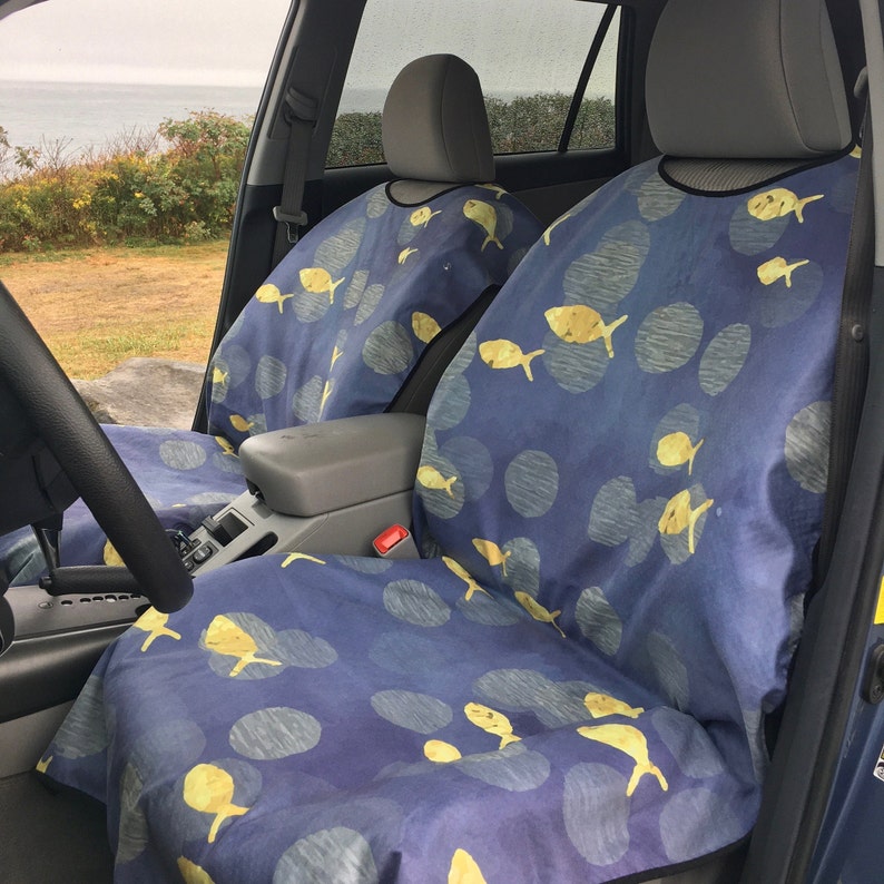 May include: A pair of car seat covers featuring a dark blue background with a repeating pattern of yellow fish and grey circular shapes. The covers are fitted over the front seats of a vehicle, extending from the headrest down to the seat cushion.