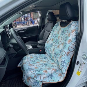May include: A light blue car seat cover featuring a repeating pattern of dark blue sea turtles, brown palm trees, and brown rectangular outlines. The cover has a tan trim and is fitted over the front passenger seat of a vehicle with black upholstery.