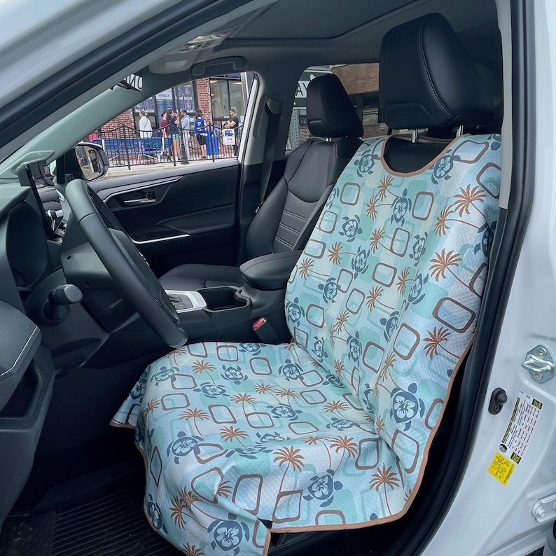 May include: A light blue car seat cover featuring a repeating pattern of dark blue sea turtles, brown palm trees, and brown rectangular outlines. The cover has a tan trim and is fitted over the front passenger seat of a vehicle with black upholstery.