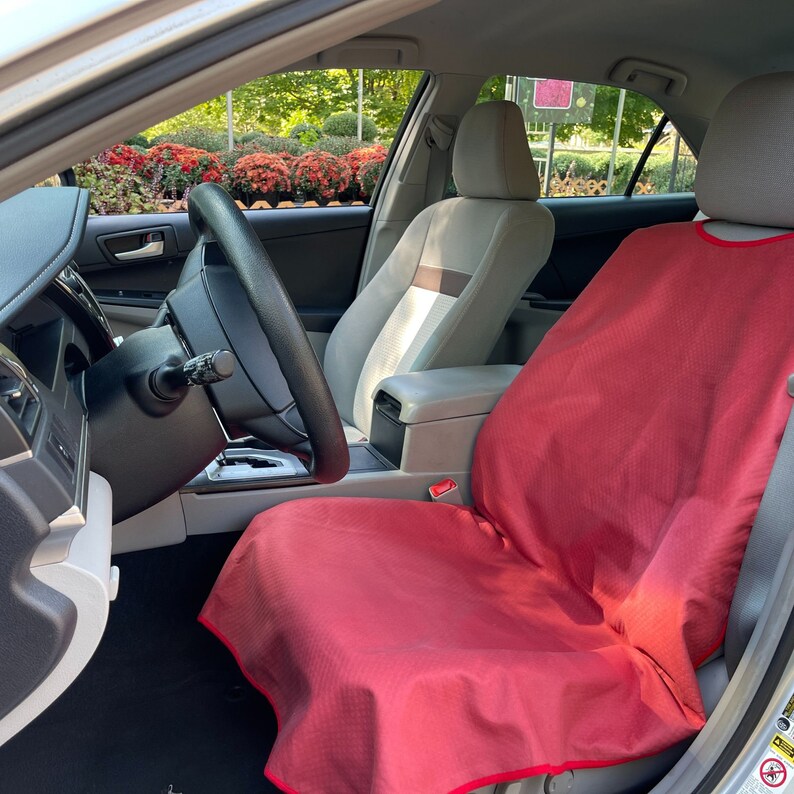 May include: A bright red car seat cover with a subtle textured pattern and a matching red trim, draped over the front passenger seat of a vehicle. The cover extends from the headrest down to the seat cushion, providing a protective layer.