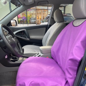 May include: A bright purple car seat cover draped over the front passenger seat of a vehicle. The cover features a smooth, solid-colored fabric that extends over the backrest and seat cushion, contrasting with the light gray interior of the car.