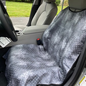 May include: A gray car seat cover featuring a metallic diamond plate pattern. The protective cover is draped over the front passenger seat of a vehicle, extending from the headrest down to the seat cushion, with black trim along the edges.