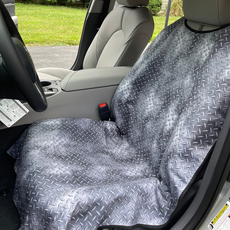 May include: A gray car seat cover featuring a metallic diamond plate pattern. The protective cover is draped over the front passenger seat of a vehicle, extending from the headrest down to the seat cushion, with black trim along the edges.