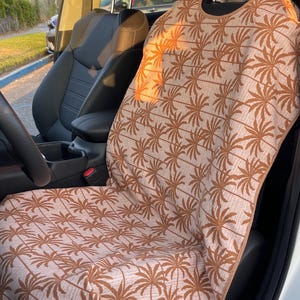 May include: A car seat cover featuring a repeating pattern of brown palm trees on a light beige fabric background. The cover is draped over the front passenger seat of a vehicle, with brown trim along the edges and a cutout for the headrest.