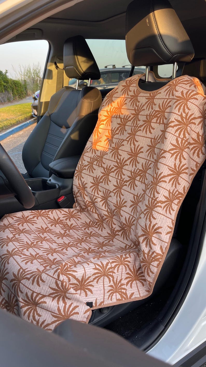 May include: A car seat cover featuring a repeating pattern of brown palm trees on a light beige fabric background. The cover is draped over the front passenger seat of a vehicle, with brown trim along the edges and a cutout for the headrest.