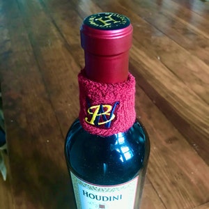 May include: A red wine bottle with a black and gold label that says "HOUDINI" and a red knit wristband with a gold and blue monogram.