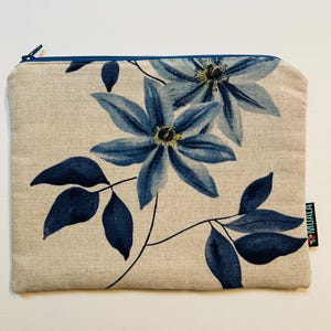 May include: A beige zippered pouch featuring a floral design with large blue flowers and dark blue leaves. The pouch has a blue zipper and a small black tag with white text and a colorful logo.