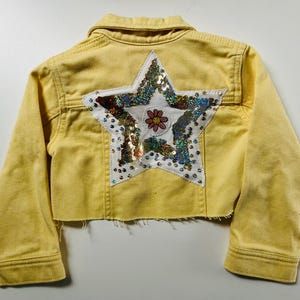 May include: A cropped yellow denim jacket. The back features a large white star, embellished with iridescent sequins and a small flower. The jacket has a classic collar and button closures. The jacket's length is cropped.
